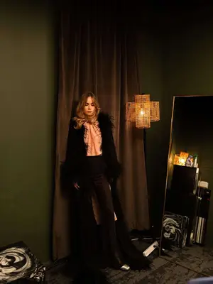 Suki Waterhouse wears a pink blouse, black skinny pants, and black feather trim coat