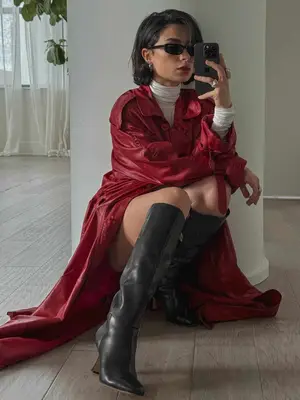 Woman takes mirror selfie while wearing red coat, white turtleneck, sunglasses, and tall black boots