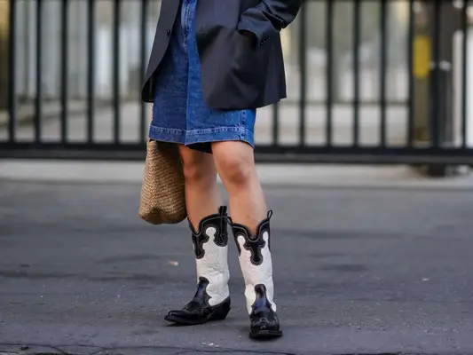 Close-up of woman wearing denim Bermuda shorts, gray blazer, woven tote bag, and black and white cowboy boots