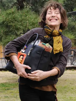 Jane Birkin wears a brown sweater, yellow scarf, and carries a black Birkin bag with charms and stickers