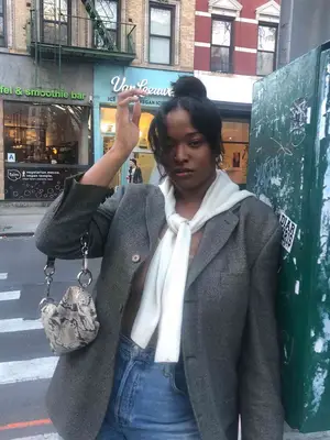 Imani Randolph wears a gray blazer, cream sweater tied over shoulders, jeans, snakeskin bag, and topknot hairstyle