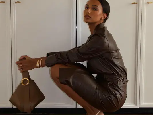 Jasmine Tookes wears a brown leather dress, layered gold bracelets, and a brown leather bag with gold details