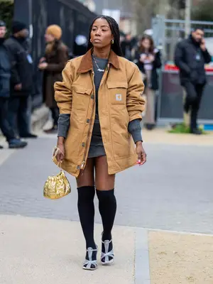Woman wears beige Carhartt barn jacket, gray sweater dress, knee-high black socks, strappy heels, and gold handbag
