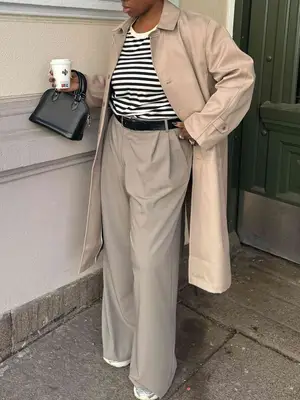 Nnenna Echem wears a black and white striped top, khaki trench coat, trousers, sneakers, and black handbag