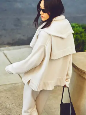 Model wears layered white cashmere sweaters, pants, a black handbag, and sunglasses