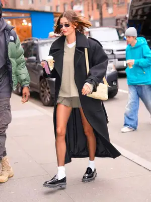 Hailey Bieber wears an oversized gray coat, beige sweater and mini skirt, black loafers with white socks, woven shoulder bag, and sunglasses