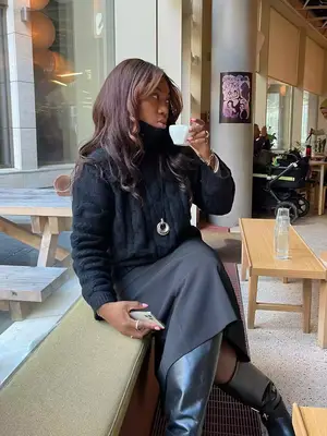 Nnenna Echem wears a black sweater, midi skirt, leather knee-high boots, and assorted jewelry