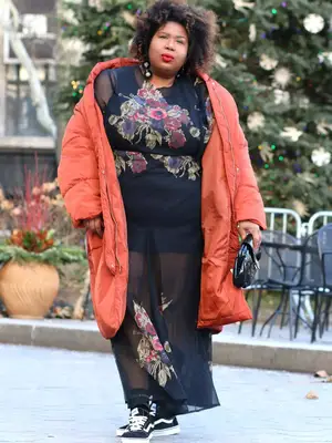 Kellie B wears a black semi-sheer dress with florals, orange coat, handbag, black sneakers, and dangling earrings