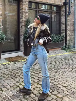 Sofia Richie Grainge wears a brown leather jacket with shearling trim, off-white sweater, light wash jeans, brown platform shoes, beanie, and green sunglasses