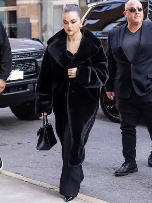 Selena Gomez wears a black fur coat, trousers, boots, and handbag