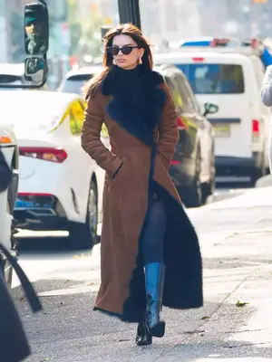 Emily Ratajkowski wears a brown and navy suede and shearling coat, turtleneck, leggings, black leather boots, sunglasses, and gold earrings