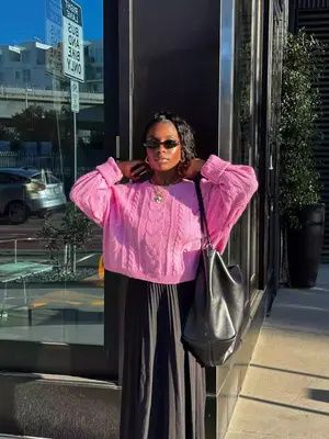 Toni Bravo wears a pink cable knit sweater, black pleated skirt, oversized black handbag, sunglasses, and gold jewelry