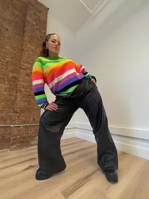 Ashley Graham wears a colorful striped sweater, black wide leg pants, platform shoes, gold earrings, and ponytail hairstyle