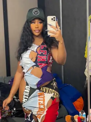 SZA taking mirror selfie wearing a green baseball cap with an upside smiley face and a cutout college t-shirt dress