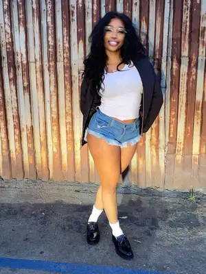 SZA wears glasses, a white tank top, a black jacket, cut off shorts, and Dr.Martens loafers.