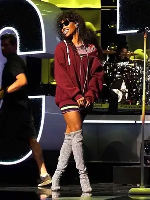 SZA wears sunglasses, a red oversized hoodie, and silver glitter boots