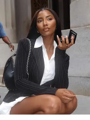 Person wearing a pinstriped blazer, white shirt dress, black handbag, and holding a mirror compact
