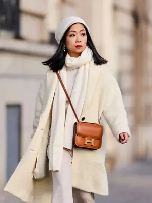 May Berthelot wears a white cashmere beanie, a matching white cashmere scarf, an off-white coat, a brown leather Hermes Constance crossbody bag, and beige flared pants 