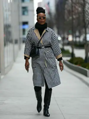 A dark skin person is wearing brown square sunglasses, silver earrings, a black ribbed wool turtleneck pullover, a black and white checkered print pattern long coat, a black shiny leather belt, a black shiny leather crossbody bag, black legging pants, black shiny leather crocodile print pattern pointed / block heels knees boots / high boot, 