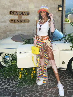 Winnie Harlow wears white tank top, orange bikini bottoms, striped crochet sarong and hat, yellow Chanel bag, and white sneakers