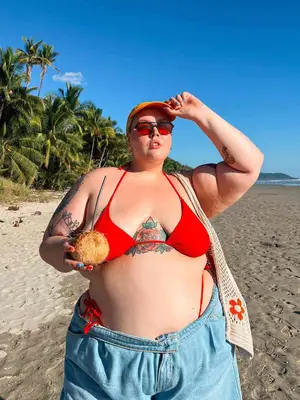 Abby Bible wears a orange baseball cap, sunglasses, a red string bikini, shorts, and a crochet handbag