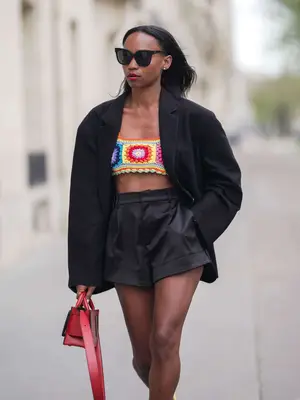  Emilie Joseph wears sunglasses, a black oversized blazer jacket by The Frankie Shop, black high waist satin shorts, bright pastel yellow leather pumps / shoes by Cosmoparis, a colored yellow and red crochet knit wool bralette /crop top with floral patterns, a red leather bag from Boyy, during a street style fashion photo session