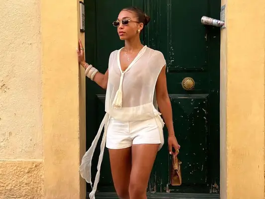 Lori Harvey wears a sheer white top, neutral bikini top, white shorts, brown handbag, and sunglasses