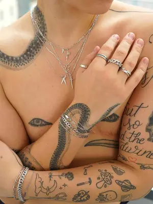 Close-up of tattooed person with top surgery scars wearing layered silver necklaces, bracelets, and rings