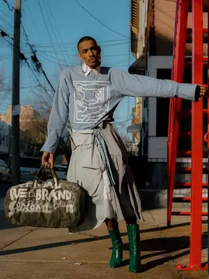 Person wearing button-down shirt with graphic design, white collar, gray skirt, graffiti duffel bag, green boots, and oversized earrings