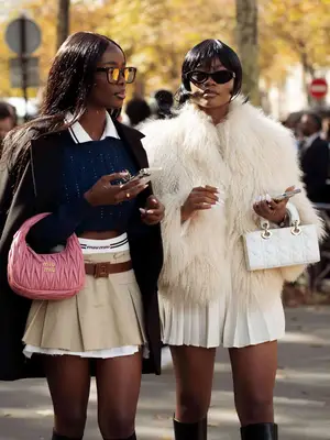 Two people pose for a photo outside. One wears a sweater, mini skirt, coat and carries a pink bag, while the other person wears a beige fur coat and pleated skirt. 