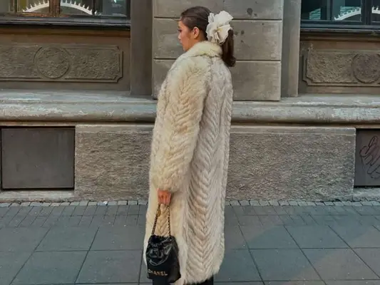 Woman wearing a large fur coat, hair accessory, and carrying a Chanel bag