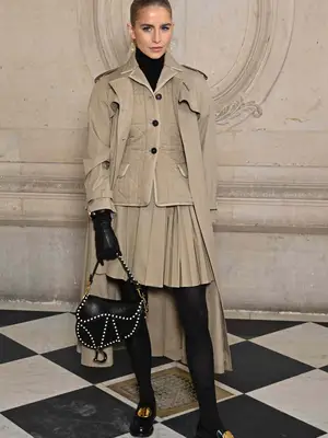 Caroline Daur posing in all khaki outfit of tench coat, vest, pleated tennis skirt, black tights, black leather gloves, dior saddle bag