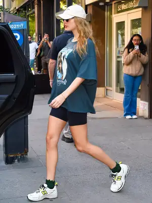 Taylor Swift wearing blue oversized tee, white hat, sunglasses, black biker shorts, and black socks popping out of New Balance white sneakers