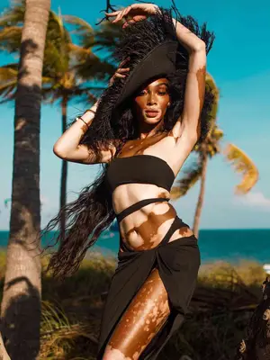 Winnie Harlow wears a black cutout swimsuit with a black sarong and large black hat with fringe