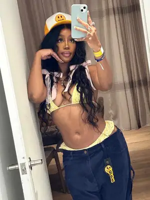 SZA wears a yellow floral bikini with dark wash, a yellow and white trucker hat with a smiley face, and pigtails with pink bows