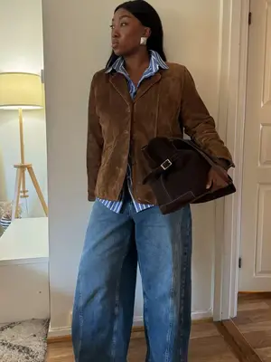 Nnenna Echem wears a blue striped button-down, brown suede jacket, and baggy mid-wash jeans. She carries a large shoulder bag. 