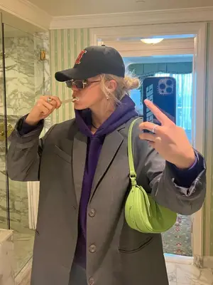 Renee Rapp brushes her teeth while wearing a gray blazer, purple hoodie, baseball cap, and lime green shoulder bag.