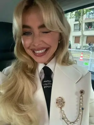 Sabrina Carpenter wearing a white blazer, collared shirt, tie, and various gold and pearl brooches