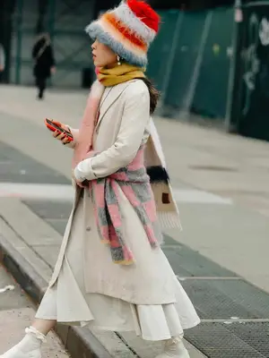 Linda Cui Zhang wears a beige structured coat, ivory skirt and ruffle socks, colorful checkered sweater tied around waist, knit colorful scarf, and fuzzy hat