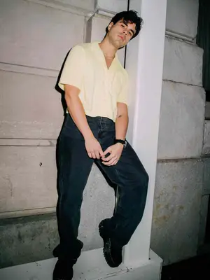 Matthew Cancel wears an off-white button-down shirt, wide leg jeans, and sneakers