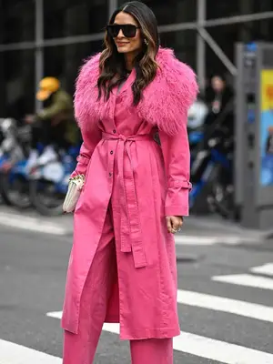 Divya Mathur wears oversized sunglasses, a pink longline coat with faux fur detailing, and pink trousers