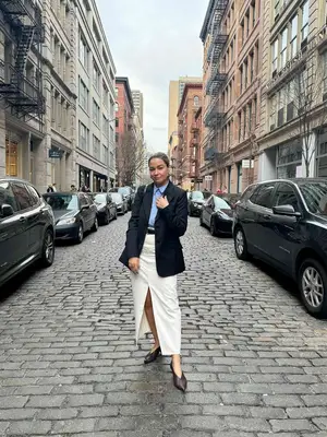 Irina Grechko wears a blue button-down shirt, white maxi skirt, black blazer, and dark pointed-toe shoes