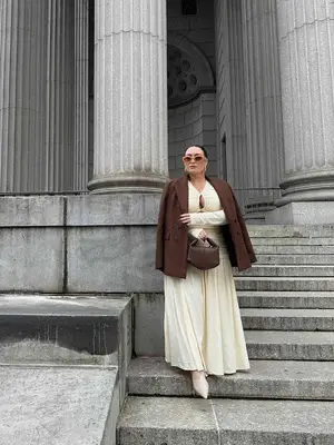 Bella Gerard wears an ivory maxi dress, off-white pointed toe shoes, brown blazer and handbag, and sunglasses