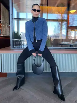 Michelle Ochs wears a denim bandage Herve Leger jacket, black turtleneck and pants, foldover boots, structured handbag, and sunglasses