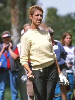 Celine Dion holding a golf club wearing yellow sweater, dark jeans, and short blonde hair cut