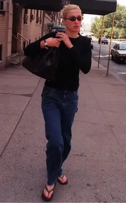 Carolyn Bessette-Kennedy wearing sunglasses, a black sweater, jeans, and flip flops