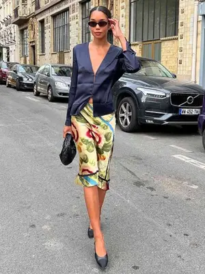 Laura Harrier wears a printed skirt, navy top, ballet flats, sunglasses, and black clutch bag