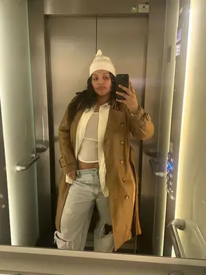 Paloma Elsesser wearing a camel trench coat, button down, sheer tank top, light-wash jeans, and a hat