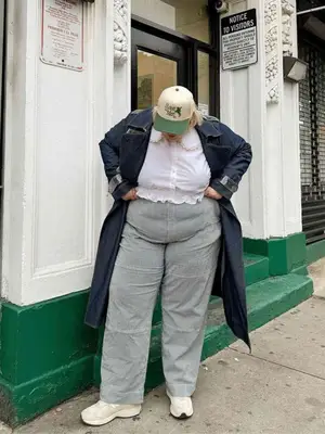 Abby Bible wearing gray pants, white blouse, denim trench coat, and a trucker hat