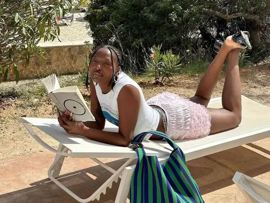 Woman wearing white graphic tank, pink bloomer shorts, and black heeled sandals while lounging on chair and reading book
